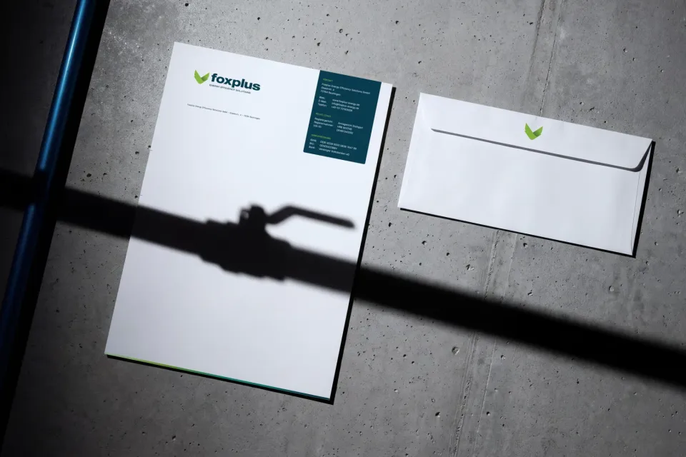 foxplus: letterhead with logo and petrol-coloured contact block next to an envelope with the mark, on concrete, with the shadow of a ball valve across it