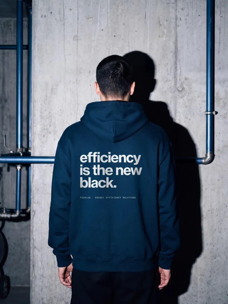foxplus: dark blue hoodie seen from behind with the print “efficiency is the new black.” and the line “foxplus · Energy Efficiency Solutions”, in front of a concrete wall with blue pipes