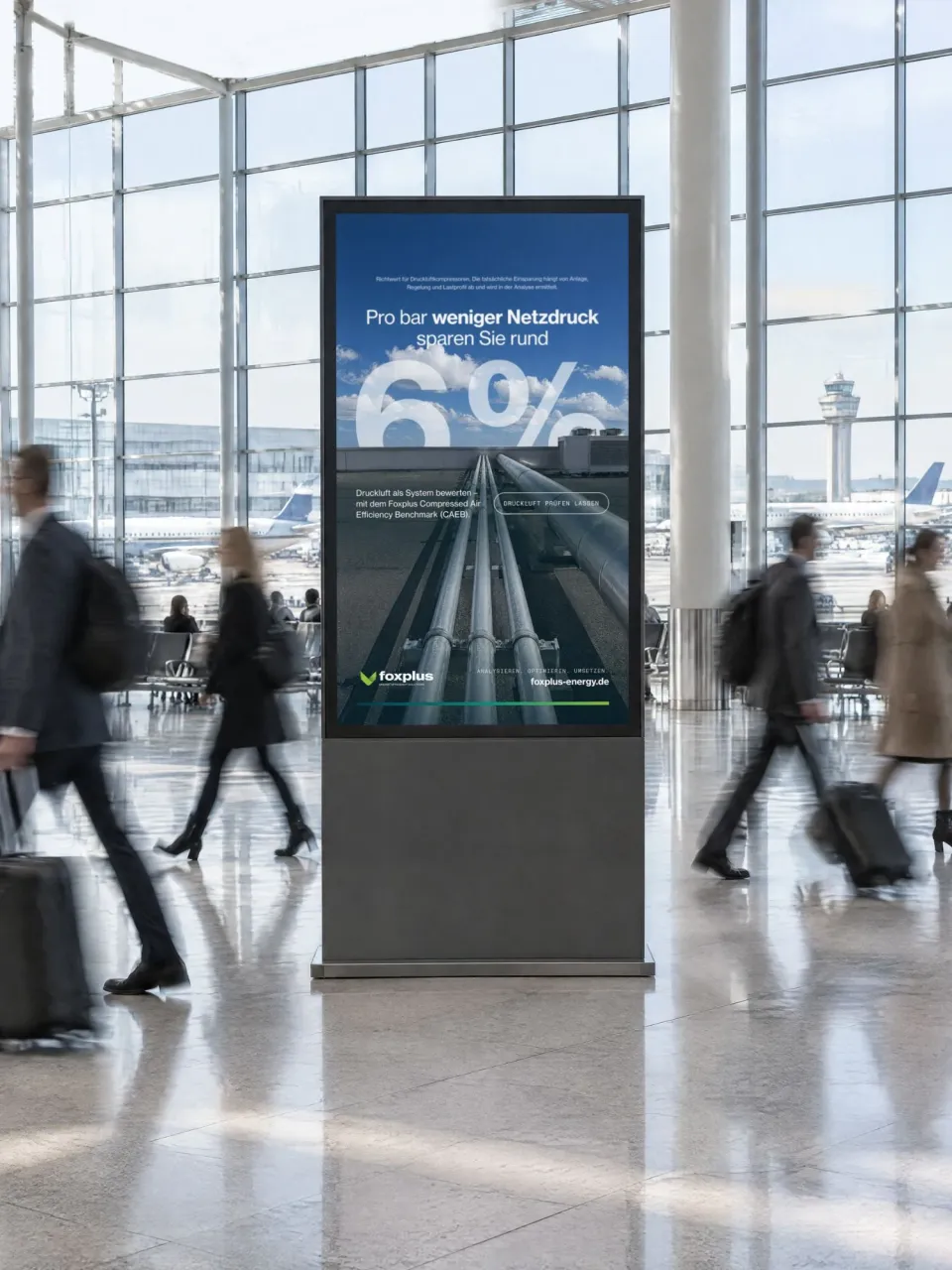 foxplus: digitale Stele in einem Flughafenterminal mit Druckluftleitungen unter blauem Himmel, der Zahl „6 %“ und der Zeile „Pro bar weniger Netzdruck sparen Sie rund 6 %“, Reisende in Bewegung