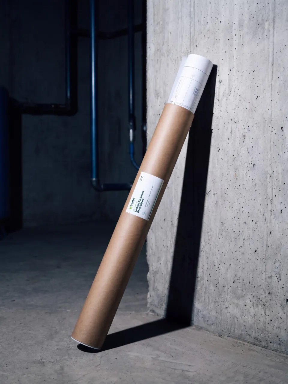 foxplus: drawing tube labelled “Leckage-Kartierung Druckluftnetz”, leaning against a concrete wall with blue compressed-air pipes behind