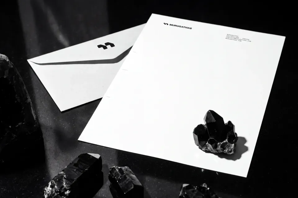 Murugathas: letterhead with mark and wordmark in the header and a contact line Deutschland · Indien, next to it an envelope with the mark, among black crystals on a dark surface, black and white
