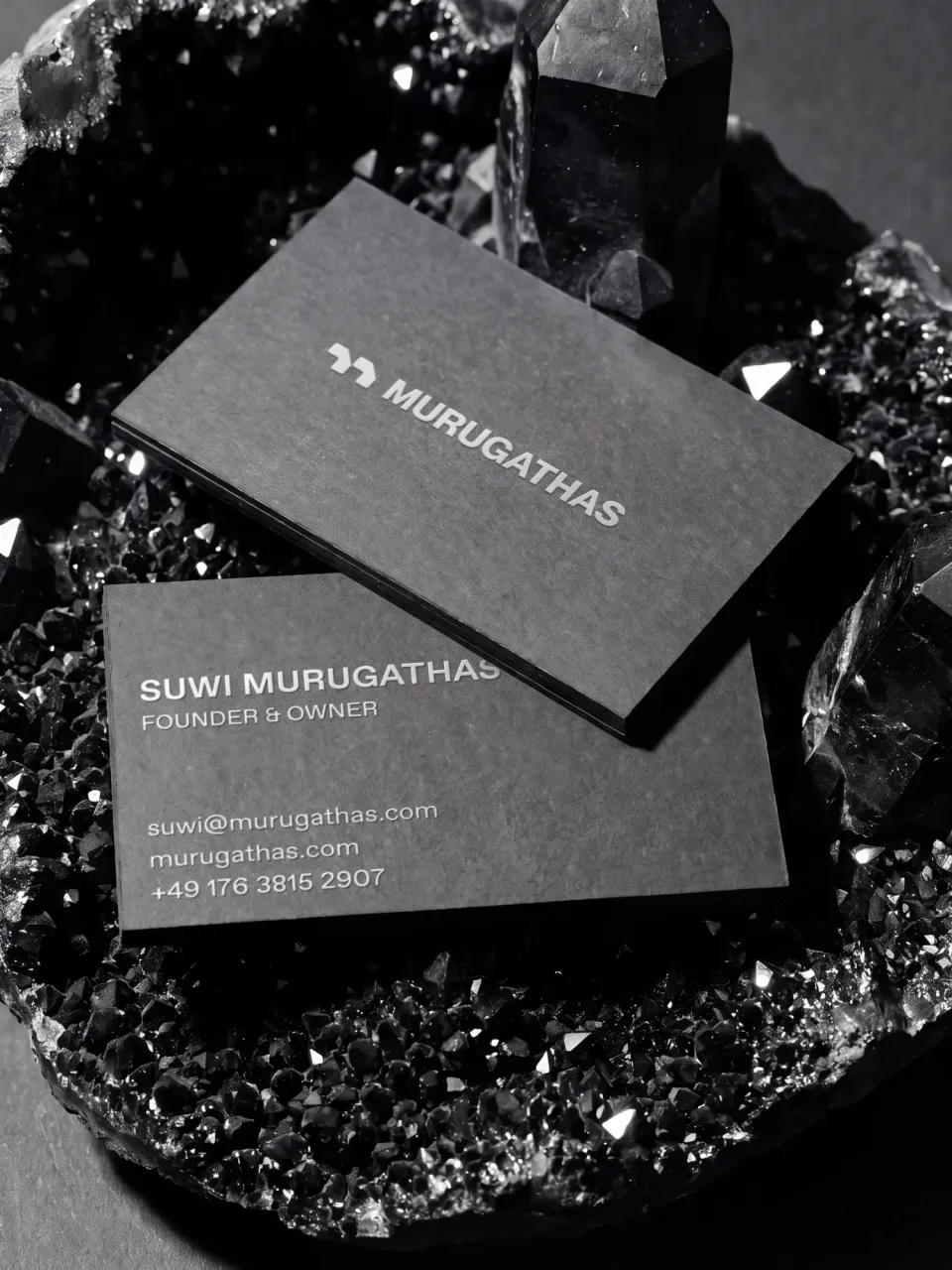 Murugathas: two black business cards with the white logo and the contact details of Suwi Murugathas, Founder & Owner, resting in a black crystal geode